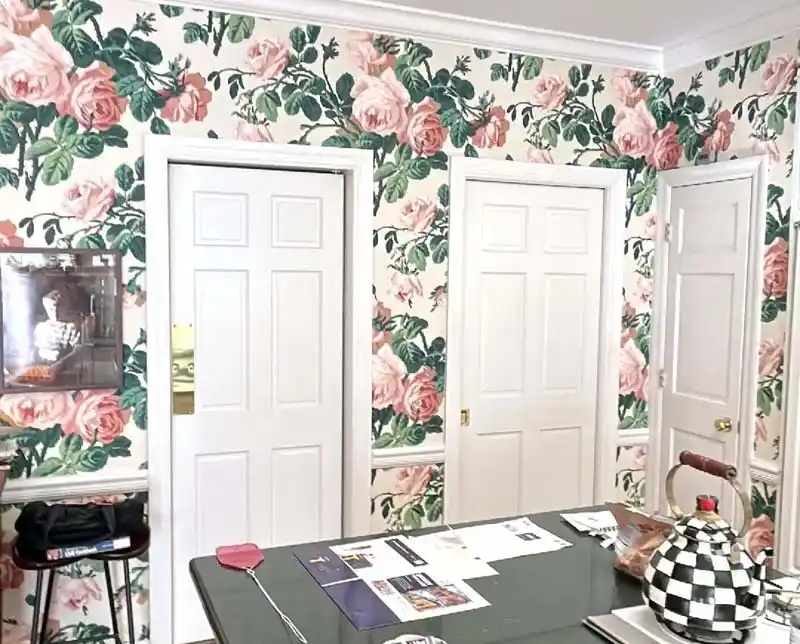 A room with three white doors and a vibrant floral wallpaper with a large rose pattern. In the foreground, there's a table with papers and a black-and-white checkered teapot. A framed picture hangs on the wall next to the first door.