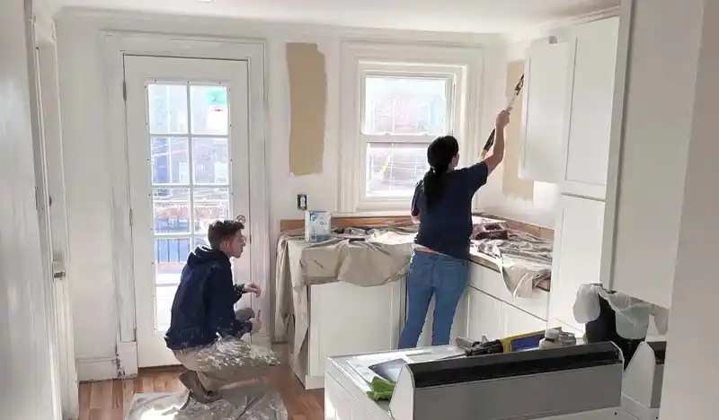 Two people are in a kitchen, painting the walls a light beige color. One person is on the right, using a roller to paint a wall section, while another person kneels on the floor to the left, working near a white door with a window. The kitchen has white cabinets and white trim.