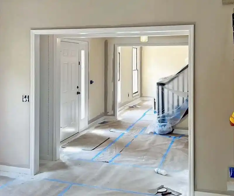 An empty entryway and staircase of a house under renovation. The floors are covered with a protective layer and blue painter's tape, and the staircase railing is wrapped in plastic. The walls are painted a light beige, and the trim and doors are white.