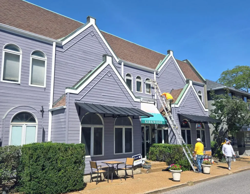 A painter on a ladder working on the exterior of a two-story gray building with multiple gabled sections. A second person is standing below the ladder, and a third person is walking away. The building has a sign that says "KIRKWOOD