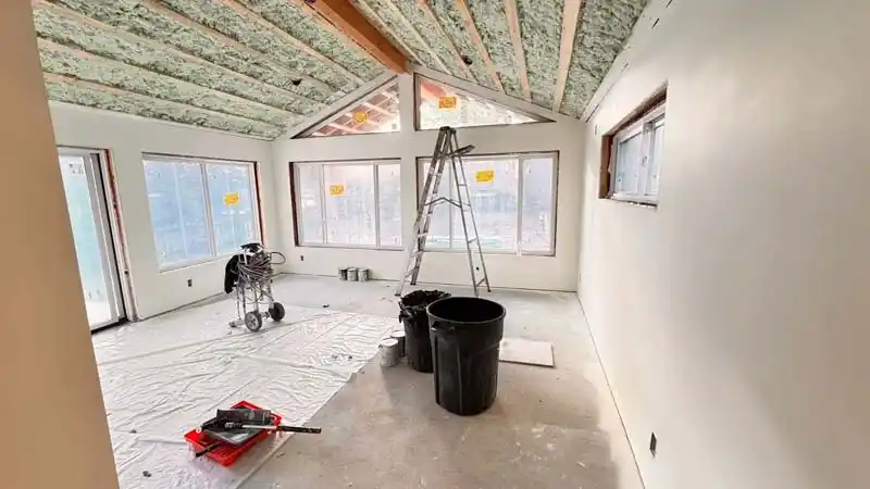 A room under renovation with white walls and an unpainted vaulted ceiling with exposed wood beams and insulation. The floor is covered in a drop cloth, and painting supplies, including a roller, ladder, and paint sprayer, are visible.