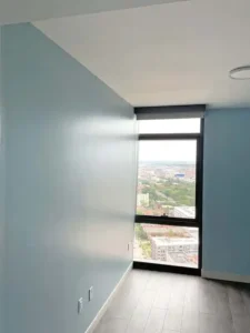 A bright room with light blue walls and a gray wood floor. A large window with a black frame and a black shade looks out onto a city with a mix of buildings and trees.