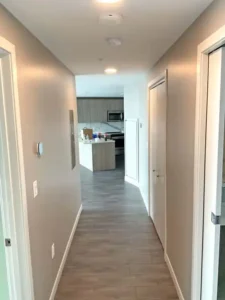 A view down a hallway with beige walls and a gray tiled floor. The hallway leads to an open-plan kitchen with light wood cabinets, a white countertop, and a microwave. There are two white doors on the right side of the hallway and a thermostat on the left.