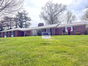 A red brick, ranch-style house with a long white section and a gray roof. The house has black shutters on the windows and two brick chimneys. Three people are standing in the front yard near a sign for "MALONE" with a phone number. The sky is overcast.