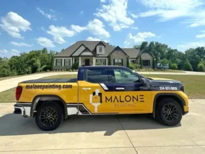 A yellow Malone Painting company truck is parked in the driveway of a large, two-story house with stone and siding on a sunny day. The truck has "malonepainting.com" and "MALONE PAINTING" on the side