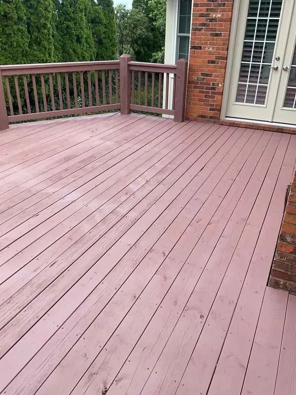 A newly finished, reddish-brown wooden deck with a wooden railing and a set of stairs leading to a lower level. The deck is attached to a red brick house with a white door and window frames. The deck boards are clean and evenly painted.