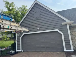 An outdoor, eye-level shot of a garage with gray siding and a gray garage door. The garage door is framed by white trim, and a small white owl figurine is mounted on the wall above the door. The top portion of the garage has a decorative louvered vent. A ladder is propped against the left side of the garage, partially obscuring a basketball hoop in the yard. The driveway is paved with red brick pavers