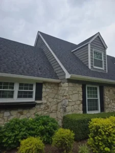 An outdoor, close-up shot of the front of a house. The house features a mix of materials, including light-colored stone on the lower portion and gray siding on a dormer and a second-story peak. The roof is covered in dark gray shingles. There are windows on the first floor with black shutters, and a window is visible in the dormer on the second floor. A low, well-maintained hedge is in the foreground. The sky is overcast