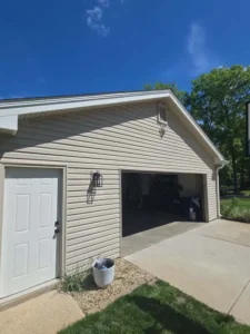 An outdoor shot of a two-car garage with light-colored siding. The garage door is open, revealing the interior. To the left of the garage door is a white pedestrian door with a window. A white bucket sits between the two doors. A black light fixture is mounted on the wall above the garage door. The sky is bright blue with a few clouds. The foreground shows a concrete driveway and a grassy area