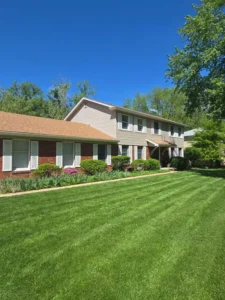 An outdoor shot of a two-story house with a large, manicured lawn in the foreground. The house features a mix of red brick on the left and light brown siding on the right. A low, well-maintained hedge runs along the front of the house. The windows have white shutters. The roof is a reddish-brown color. The sky is clear and bright blue. Trees and greenery are visible in the background