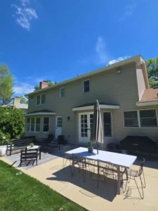 An outdoor shot of the back of a two-story house with tan siding. The house has a two-tiered patio area. The patio in the foreground has a white table with wireframe chairs and a large gray umbrella. The patio area closest to the house has a dark wicker bench, chairs, and a small table on a patterned rug. A white door with glass panels leads into the house. A bay window is on the left side of the house. The yard is grassy, and the sky is bright blue with some wispy clouds