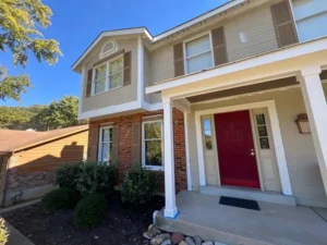 The front of a two-story house with a red door and a covered porch. The house has a mix of red brick and light-colored siding. There are bushes in front of the house and a brown garage is visible to the left. The sky is blue with no clouds