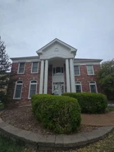 A grand, two-story house with a red brick facade is shown from a low-angle perspective. The house features a prominent portico with two large, round white pillars supporting a triangular pediment. A second-story door or window with a curved top is located behind the pillars. A ladder is set up in front of the door, and a person is visible on the ladder. The windows on the house have white frames. A raised garden bed with a low stone wall is in the foreground, filled with hedges and dirt. The sky is overcast