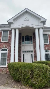 An outdoor, head-on shot of the front entrance of a two-story red brick house. A large portico with two prominent white pillars frames the main entrance. The front door is white with side windows, and a multi-paned window is located directly above it on the second story. The portico has a white triangular pediment with a decorative circle in the center. A low, manicured hedge is in the foreground, partially obscuring the base of the house. The sky is overcast and gray