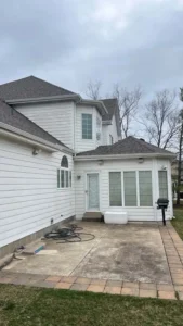 An outdoor shot of the back of a house with white siding. A small sunroom with several windows and a dark roof extends from the main house. A patio made of concrete slabs and brick pavers is in the foreground. A white storage bench sits on the patio near the sunroom door. A grill is visible on the far right. A garden hose is coiled on the patio. The sky is gray and overcast, and the surrounding trees are bare