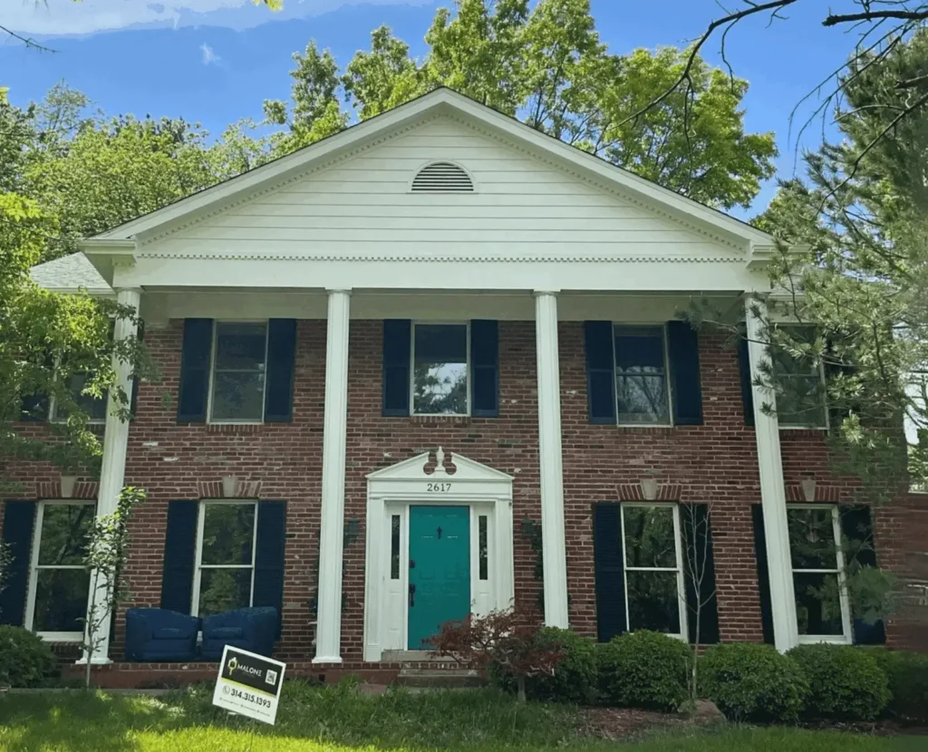 A two-story red brick house with a prominent white portico and four large white columns. The house has a bright turquoise front door, black shutters, and a white gable with a semicircular vent. A white real estate sign is visible in the front yard.