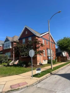 A red brick two-story house with a blue stripe running horizontally across the building. The house has a front porch with stairs, a well-maintained green lawn, and a light pole on the curb. A real estate sign is visible in the foreground.