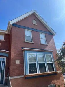 A two-story red brick house with a distinctive blue trim. The house has a large bay window on the first floor with white blinds and white-framed windows on the second floor. A horizontal blue stripe runs across the upper half of the house, and a small, arched vent is visible on the gable.