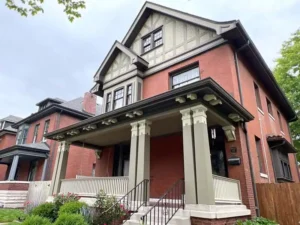A two-story red brick house with a decorative gable and a wide front porch. The porch is supported by two large gray columns and has a white railing and black metal stairs. The upper part of the house is beige with a dark brown crisscross pattern. A neighboring house is visible to the left.