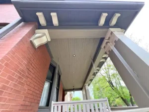 A detailed, upward-angled view of a red brick house's front porch. The porch ceiling is painted a beige and brown, with white decorative brackets. A porch swing with chains hangs from the ceiling, and a large support column stands on the right.