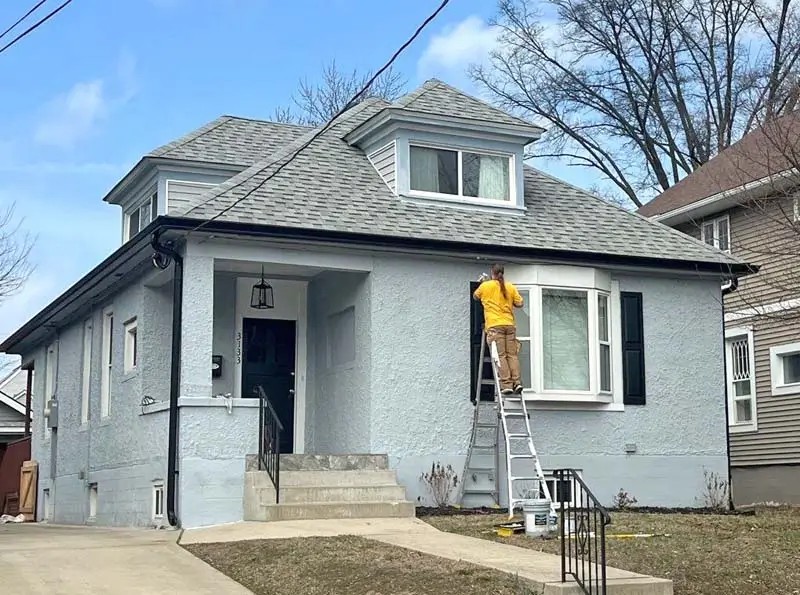 A person on a ladder is painting the exterior of a light gray house. The house has a gray shingled roof, black shutters, and a bay window. The sky is partly cloudy, and leafless trees are in the background.