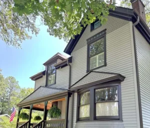 A large, light gray house with black trim and a dark, shingled roof. The house has a long porch with hanging plants and a small American flag. An arched doorway is visible on the first floor, and the lawn is green with trees and hedges. The sky is bright and blue.