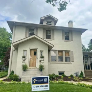 A two-story, light beige brick house with a brown front door. The house has beige shutters on the windows, a small front porch, and a dormer window on the top floor. A sign for "MALONE" with a phone number and a QR code is on the front lawn. The sky is partly cloudy.