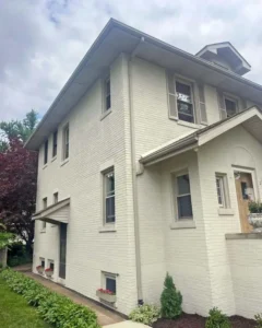 A close-up, angled view of a two-story house with a light beige brick exterior. The house has beige shutters on one window and small flower boxes below some of the windows on the side. The roof has gutters, and the sky is overcast.