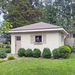 A small, light beige, one-story building with a light gray shingled roof. The building has a door, a window with light gray shutters, and a flower box. A row of neatly trimmed bushes is in front of the building, and a lush green lawn and trees surround the area.