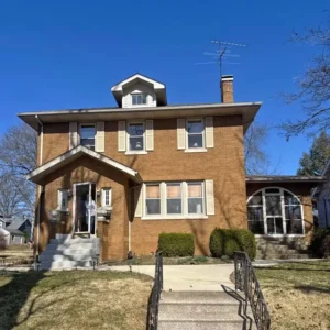 A two-story brown brick house with a small front porch and a dormer window on the roof. The house has beige shutters on some of the windows and a large arched window on the right side. A clear blue sky is visible above the house, and the lawn is dry and brown.