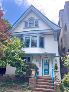 A three-story white house with blue trim and a dark gray roof. The house has a covered front porch with a blue door and a small set of brick stairs with blue metal railings. There are two large windows on the second floor and a small arched window on the third floor. Two large planters sit on either side of the bottom of the stairs.