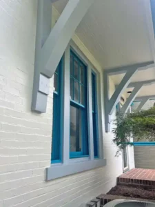 A low-angle side view of a white painted brick house. The windows have bold teal frames and gray trim. A white porch roof, supported by gray brackets, extends over the front walkway. The walkway is made of red bricks, and the house number 4460 is visible on a white sign on the far right.