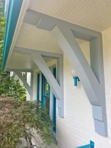 A white brick house with a front porch. The porch is supported by two large, light gray corbels and has a light fixture with a blue base. The front door and window frames are painted a bright turquoise, and a plant is visible in the foreground.