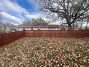 A wide view of a backyard with a newly stained red wooden fence. The ground is covered in brown leaves, and a large tree with bare branches is in the background. A small, one-story house is visible behind the fence. The sky is overcast.