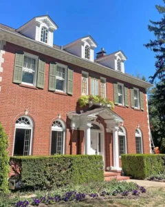 A close-up, angled view of a two-story, red brick house with a front porch. The house has green shutters on the upper-level windows and several dormer windows on the white roof. A neatly trimmed hedge is in front of the house, and a garden with purple flowers is in the foreground. The sky is a clear blue.