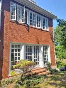 A close-up of a two-story, red brick house with a prominent bank of white-framed windows on both floors. The second-story windows are partially open. Two large columns are visible on the first floor. A small tree and a potted plant are in front of the house, and the sky is clear and blue.