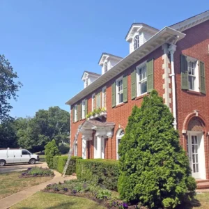 A large, red brick house with green shutters and white trim. The house has several dormer windows and a front porch with white columns. A ladder is leaning against the house, and a white van is parked on the street. The front yard has a hedge and a garden with purple flowers. The sky is a clear blue.