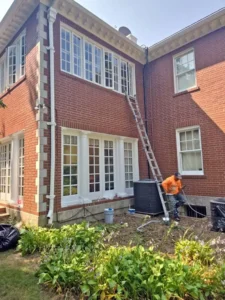 A two-story, red brick house with a white trim and white window frames. A man in an orange shirt and a baseball cap is on a ladder, working on the side of the house. An air conditioning unit is below the ladder, and a garden with green plants is in the foreground. The sky is bright and clear.