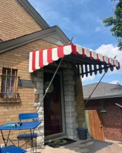 A brown brick house with a red and white striped awning over the front door. The house has a small, light-colored stone patio with a blue table and chairs. A black trash can is next to the door, and the sky is blue with some clouds.