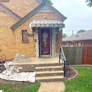 A one-story brick house with a front porch. The porch is made of concrete with three steps leading up to the front door. The front door is red with a white "Welcome" sign and is framed by stacked stone and a corrugated awning. The house number 5041 is visible next to the door. A metal handrail runs down the right side of the steps, and a wooden fence is in the background.