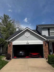 The front of a two-car garage with the door open, revealing two sports cars inside. The garage door is topped by light gray siding and a white circular vent. The lower part of the house is red brick