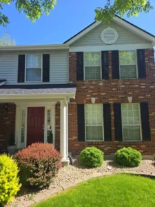 The front of a two-story brick and siding house with a red front door, covered porch, and black shutters on the windows. There are bushes and landscaping in the front yard. The sky is blue with some clouds