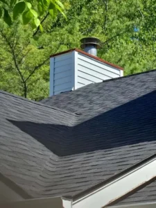 A close-up of a house roof with dark shingles and a light gray siding chimney with a metal cap. There are green trees in the background and a blue sky