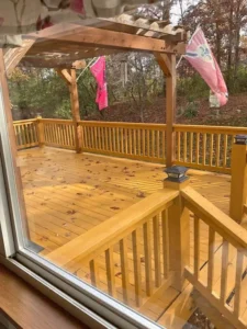 A view of a newly stained wooden deck from inside a house, looking out a sliding glass door. The deck has a natural wood color, with railings, and a small pergola overhead. A few brown leaves are scattered across the deck. Two flags are hanging from the pergola, and the deck overlooks a wooded area with trees.
