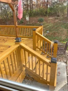 A newly stained wooden deck is visible from a large glass window, with stairs leading down to a rocky area. The deck and railings are a warm, natural wood color, and a few scattered leaves are on the surface. Solar lights are placed on the top of the newel posts, and a table with chairs is visible on the lawn beyond the deck. The background shows a wooded area with trees and dead leaves on the ground.