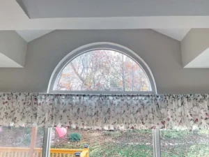 A wide-angle, interior view of a room with a large, half-circle arched window at the top, and a lower rectangular window. The windows look out onto a backyard with a deck, trees, and brown leaves on the ground. A patterned valance with a floral design hangs over the bottom window.
