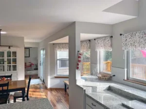 An interior view of a bright kitchen and dining area. The kitchen features granite countertops, white cabinets, and large windows with floral-patterned valances. A dining table with dark chairs is visible on the left, and a sliding glass door leads to an outdoor deck. The floors are a warm hardwood.