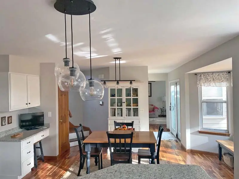 A bright, modern open-plan kitchen and dining area. The dining area features a wooden table with black chairs and a white hutch, illuminated by a multi-bulb pendant light fixture with clear glass globes. The kitchen area on the left has white cabinets, a gray countertop, and a small computer monitor. A large window with a valance is on the right.
