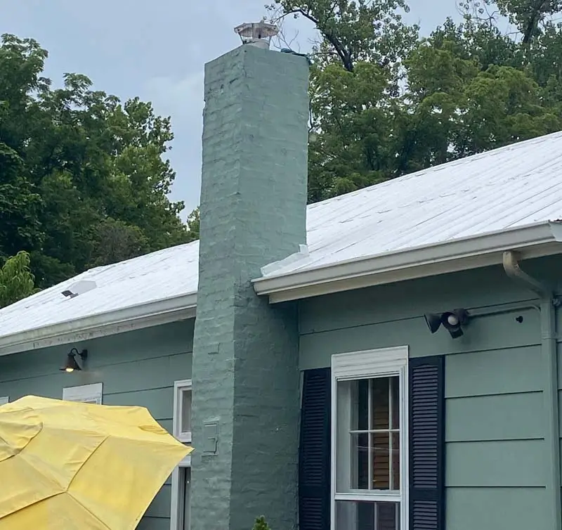A tall, mint-green brick chimney rises from the roof of a house with light green siding. The house has a white metal roof and dark shutters on the window. A yellow umbrella is visible on the left, and the sky is overcast.