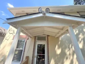 A front entrance with a small white porch roof supported by two square posts. The house has light yellow siding and red brick accents. A glass-paneled storm door is visible, and two floodlights are mounted on the roof peak above the door.